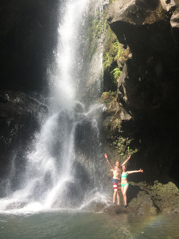 Two people in swimwear stand joyfully with arms outstretched on rocks at the base of a waterfall during their private tour in Maui. The water cascades down a rocky cliff surrounded by lush greenery, creating misty spray around them.