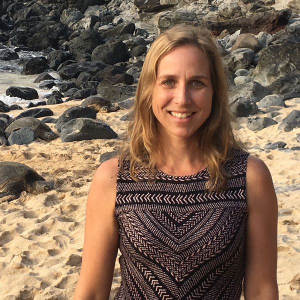 Susan on a sandy rocky beach in brown tank top with a turtle in background.