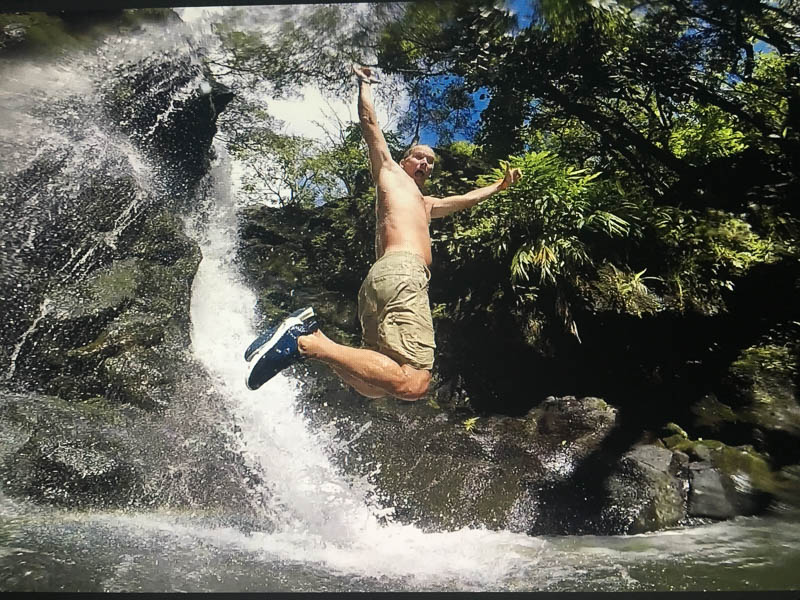A person wearing shorts and sneakers is mid-air, jumping excitedly in front of a waterfall on a Maui adventure. Surrounded by lush greenery, they have one arm extended upwards and the other along their side, capturing the thrill of the moment.