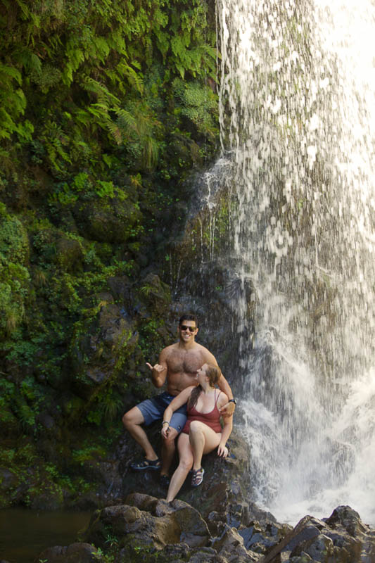 A man and woman sit on rocks beside a cascading waterfall in a lush, green setting. On this Maui adventure, the shirtless man in sunglasses smiles while gesturing with one hand. The woman, in a swimsuit, looks at him with a smile.