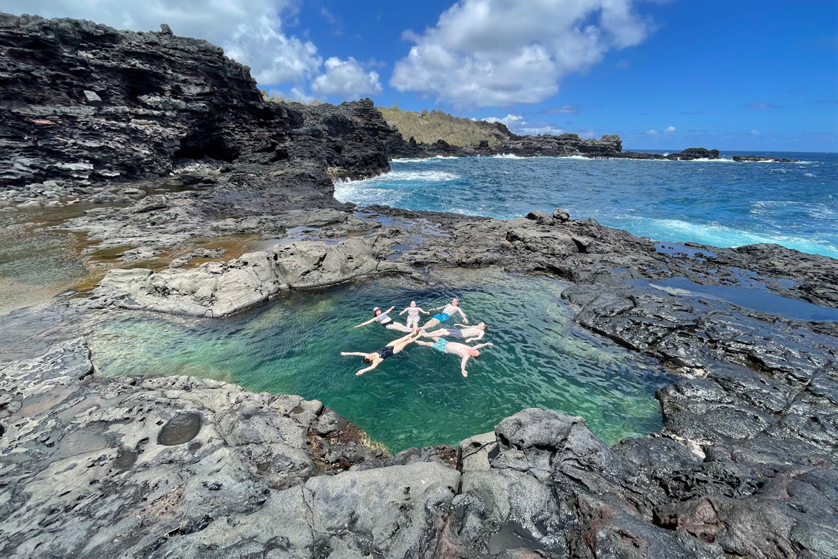 Five people float in a clear natural tide pool surrounded by black volcanic rocks, experiencing the serenity of Maui. Ocean waves crash nearby under a bright blue sky dotted with clouds, creating a picturesque moment on their private tour.