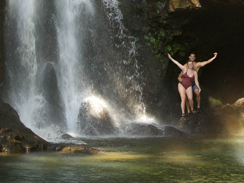 Two people in swimsuits stand on rocks under a mesmerizing Maui waterfall, arms raised in excitement. The cascade flows into a clear pool surrounded by lush greenery, creating the perfect moment on their private tour.