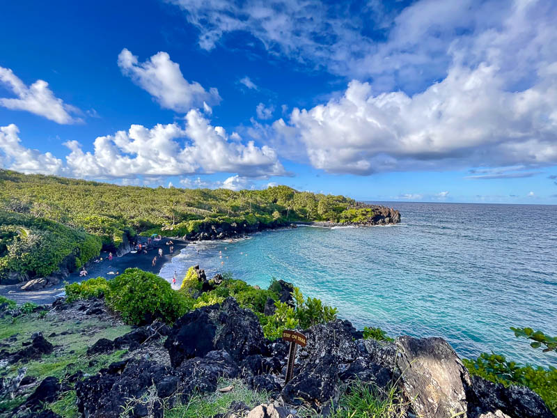 A scenic view of Maui's black sand beach with clear blue water, surrounded by lush green vegetation. People on a custom tour are enjoying the beach, and the sky is partly cloudy, creating a picturesque, peaceful setting.