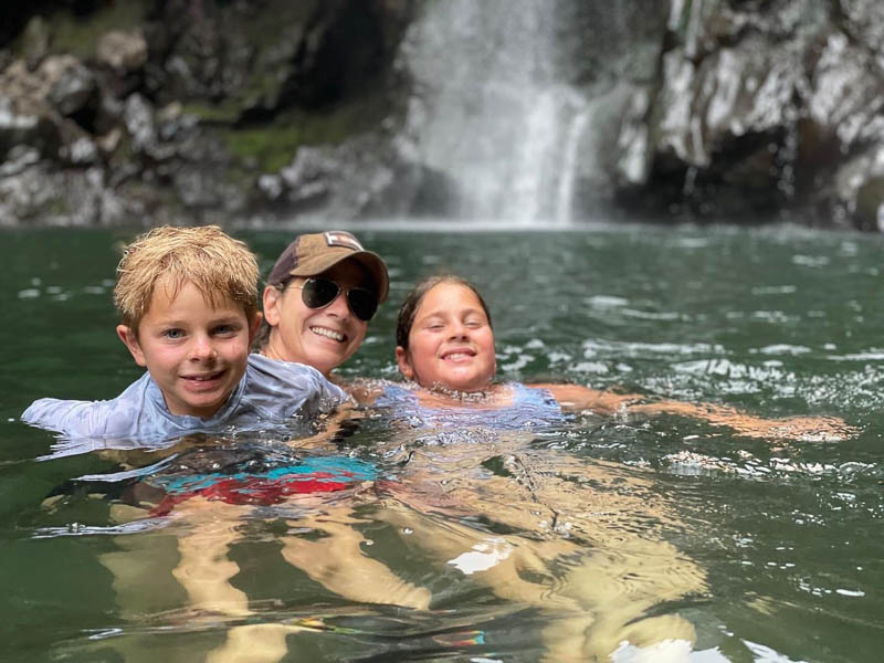 Three people swimming in a clear green pool at the base of a waterfall, enjoyed as part of a private Maui tour. Two children, one with blonde hair, and an adult wearing sunglasses and a cap, are smiling amidst the refreshing water and scenic rocky cliffs.