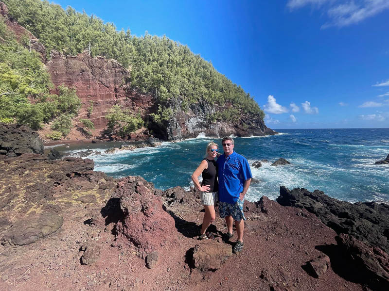 A couple poses on rugged, rocky terrain with the ocean in the background, capturing the essence of their private Maui tour. The sky is clear, and lush green cliffs are visible in the distance as they enjoy their casual summer attire.