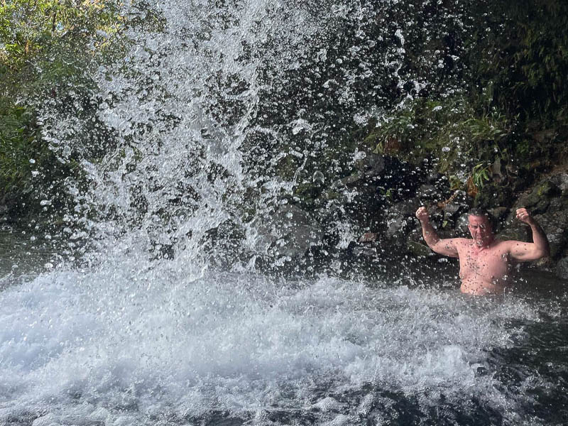 A person stands in a waterfall on a Maui adventure, arms flexed. Water splashes all around, creating a misty effect. They are surrounded by lush greenery and visible rocks, capturing the power and refreshing feel of nature during this custom tour.