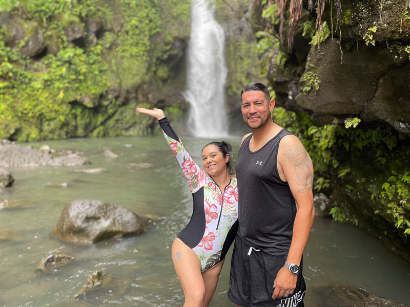 A smiling woman in a floral swimsuit stands beside a man in a black tank top and shorts, enjoying their private tour. They pose in front of a Maui waterfall surrounded by lush greenery as she raises her hand toward the cascading waters.