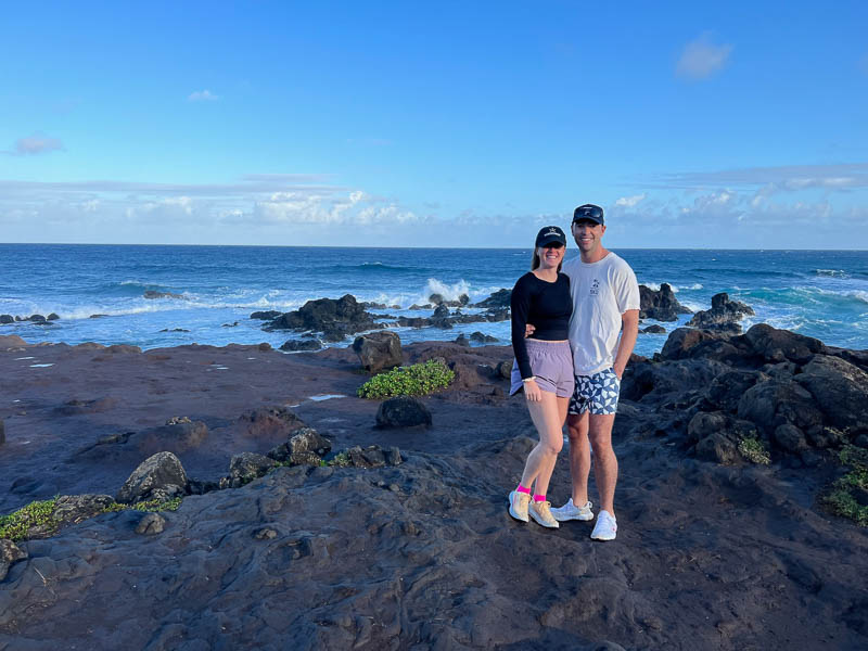 A couple stands on a rocky shoreline by the ocean under a clear blue Maui sky. The woman wears a black top and pink shorts, and the man sports a white shirt with blue patterned shorts. Both wear caps and sneakers. Waves crash on rocks in this unforgettable adventure backdrop.
