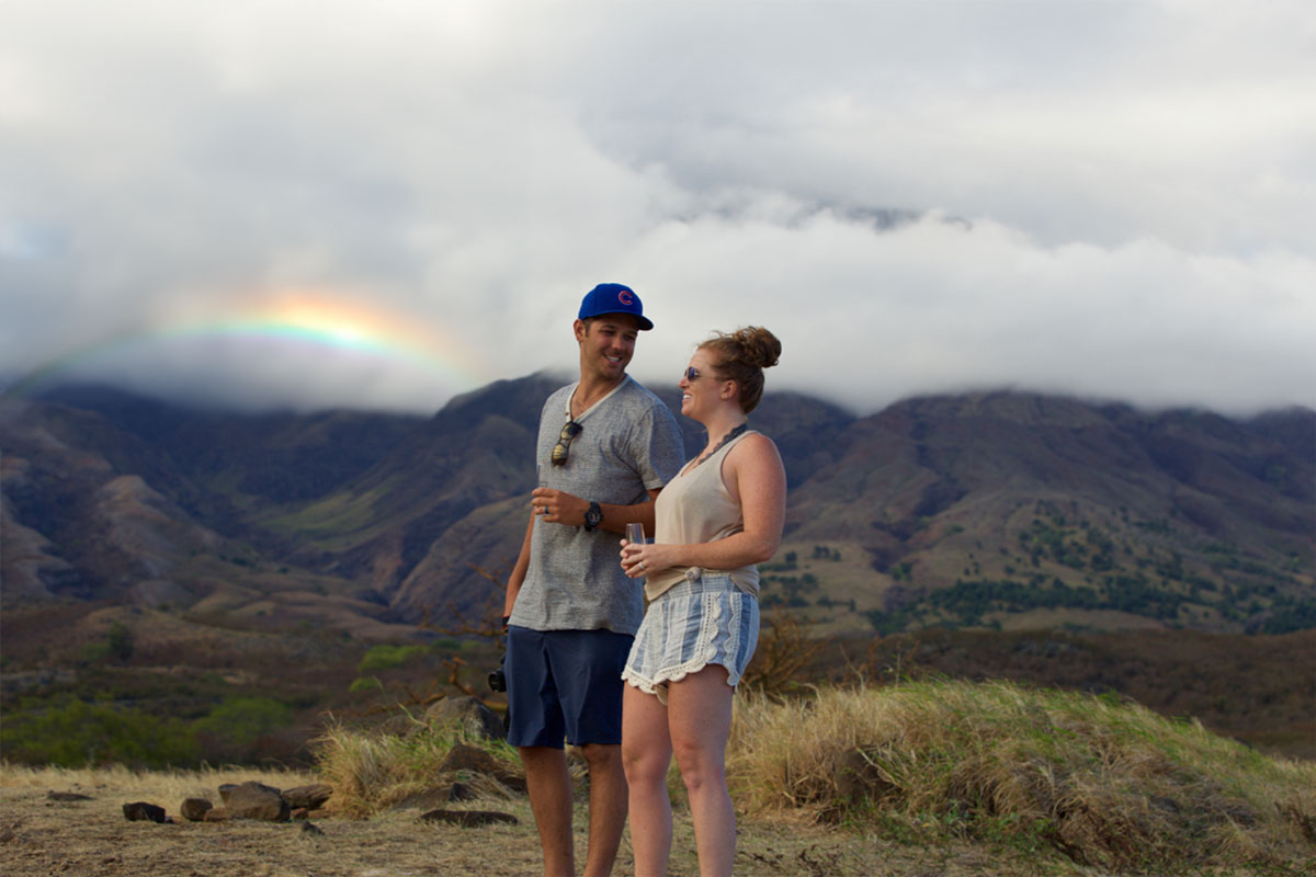 A couple stands on a hilltop with dry grass, sharing a moment, embodying the spirit of adventure. Casually dressed, the man wears a cap and holds a drink, as a picturesque rainbow arches over cloudy mountains in the background under a cloudy sky.