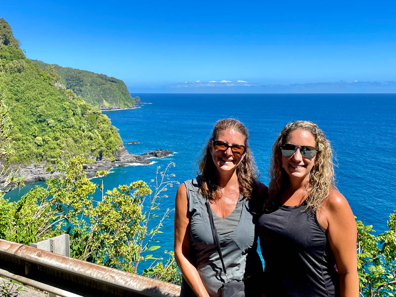 Two people stand smiling in front of a scenic coastal view, embodying the spirit of adventure. Lush green cliffs drop into a vibrant blue ocean under a clear sky. Vegetation surrounds the foreground, highlighting the natural beauty reminiscent of an unforgettable Maui tour.