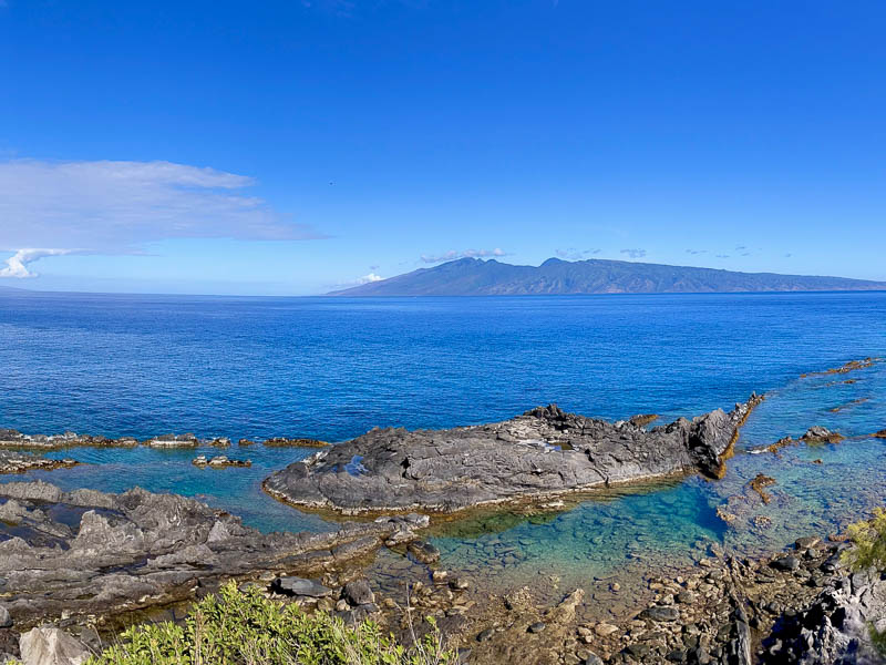 Experience a panoramic view of a rocky coastline with clear turquoise waters under a bright blue sky on a private Maui tour. In the distance, admire a large island with mountainous terrain surrounded by the vast ocean, while sparse greenery graces the foreground.