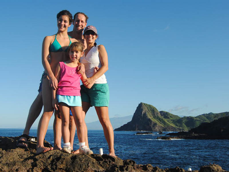 A family of four stands on a rocky Maui shoreline with the ocean and a green, hilly landscape in the background. The group is smiling and dressed in casual, summer clothing, enjoying a sunny day by the sea during their private tour.