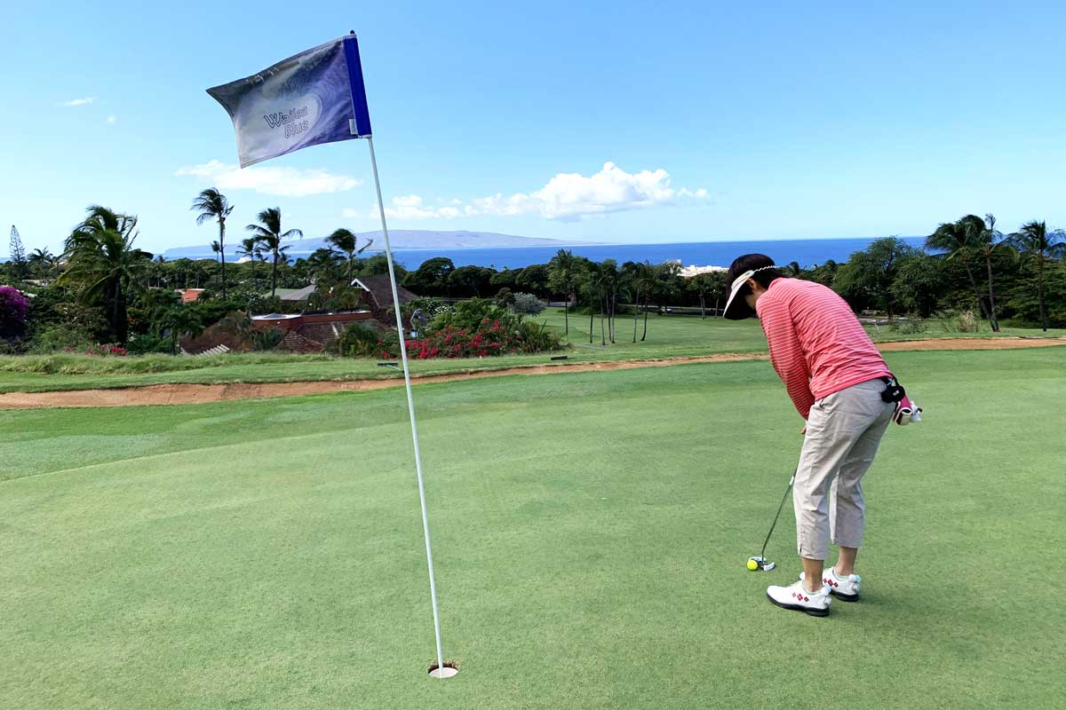 A person in a pink shirt and white hat putts a golf ball on a lush Maui green with a flag nearby. The background offers an adventurous view of palm trees, distant hills, and the blue ocean under a clear sky.