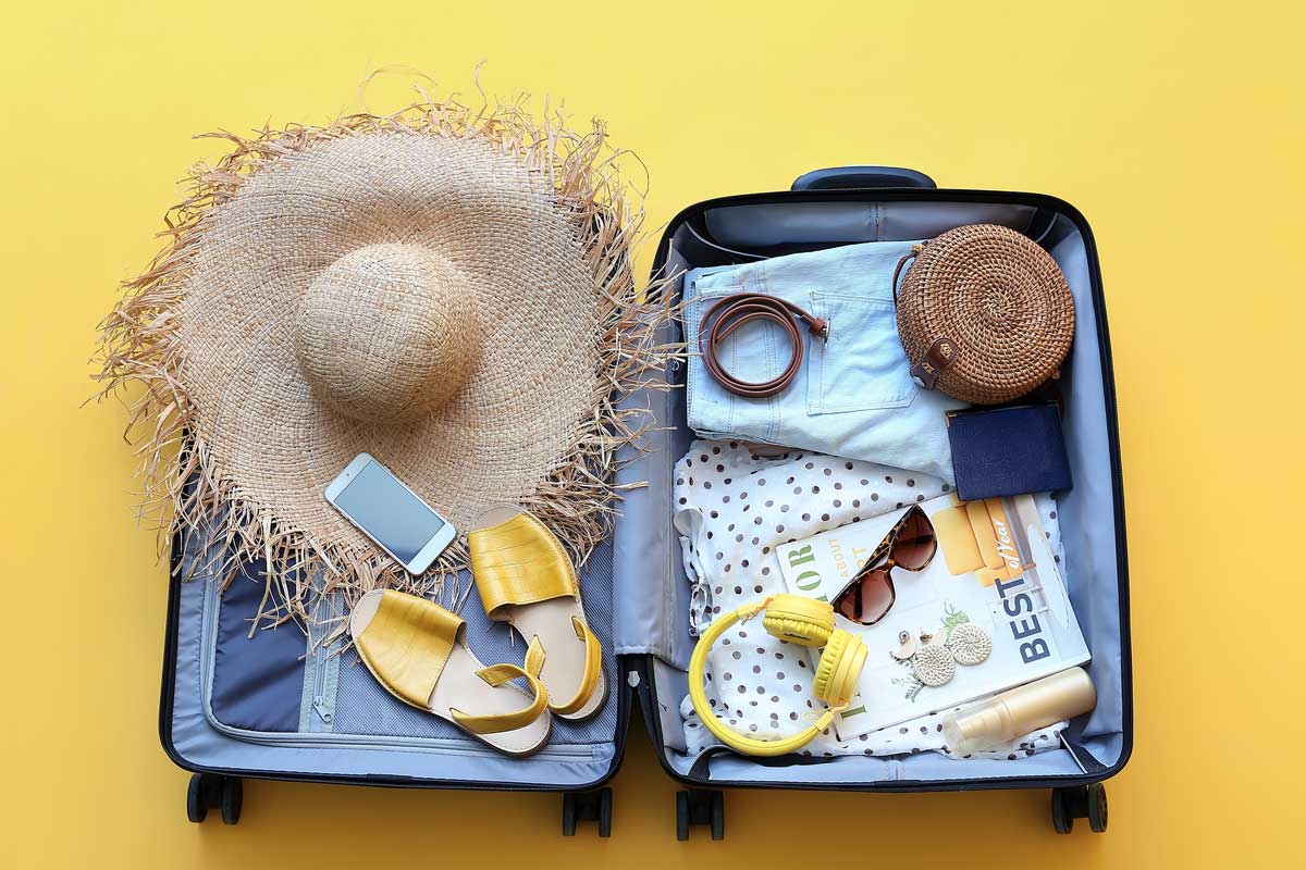 Open suitcase with a straw hat, smartphone, yellow sandals, clothes, a woven bag, headphones, sunscreen, a watch, and a magazine on a yellow background—ready for your custom tour adventure.