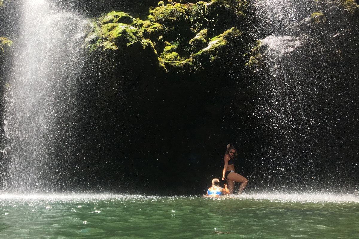 A person in a swimsuit stands under a waterfall, holding a small child in a float. Sunlight streams through, illuminating the green moss on the rocks above and casting light on the water below—a serene moment captured during their Maui tour.