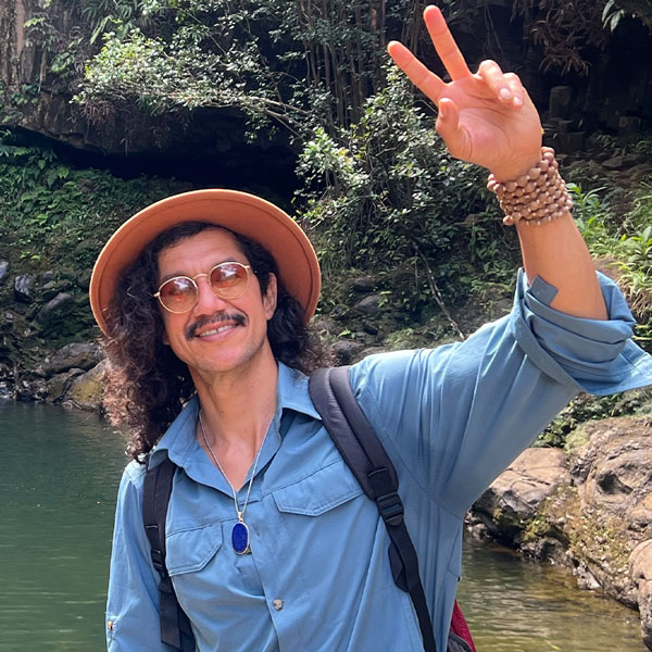 A man with curly hair, wearing a tan hat, glasses, and a blue shirt, stands outdoors by a rocky Maui stream, smiling and making a peace sign with his right hand. He has a backpack and beaded bracelets on his wrist.