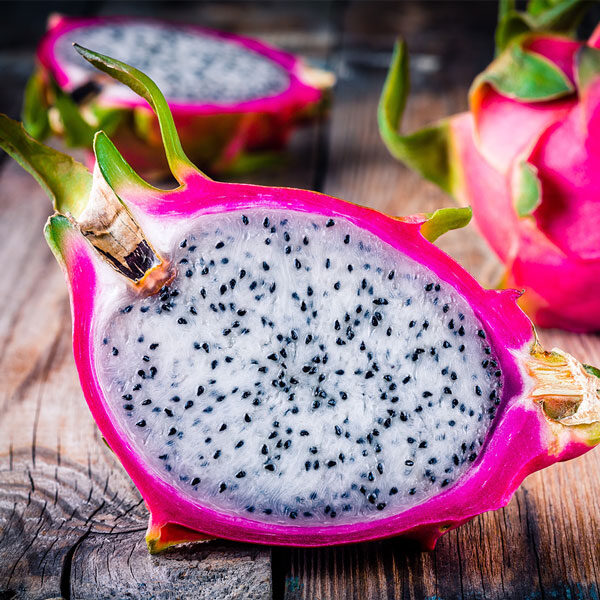 A sliced dragon fruit with vibrant pink skin and speckled white flesh rests on a wooden surface, evoking the exotic allure of a Maui tour. Another whole dragon fruit, reminiscent of luscious island adventures, is visible in the background.