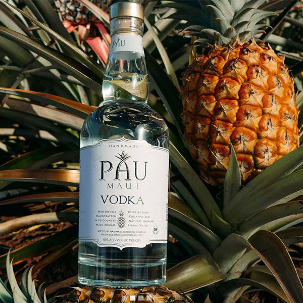 A bottle of PAU Maui Vodka is elegantly showcased among several pineapples and lush green leaves. The clear glass bottle, with its white label proudly listing the alcohol content, basks in sunlit glory. This scene perfectly captures the tropical ambiance of a custom tour through Maui's vibrant spirit.