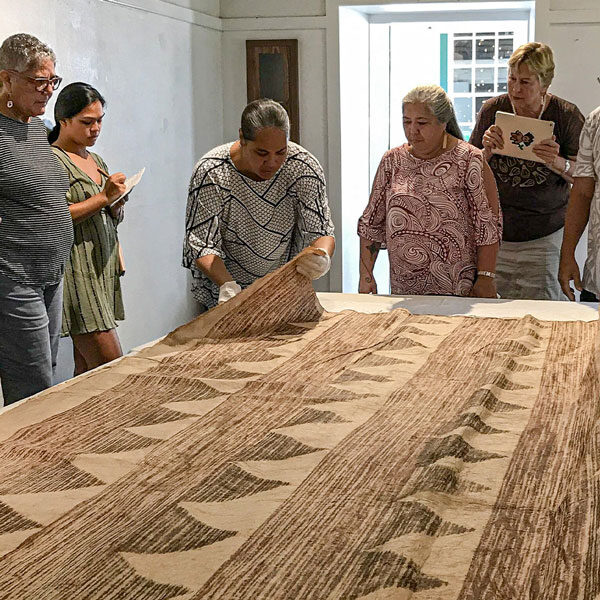 A group of people stand around a table on a custom tour, examining a piece of Tongan barkcloth, also known as tapa. One person gently lifts the cloth with gloved hands. The setting appears to be indoors with simple white walls, offering an intimate glimpse into art conservation.