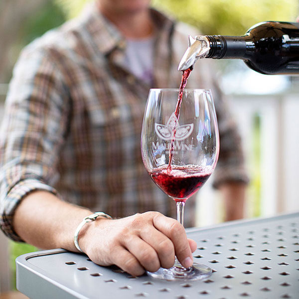 A person in a plaid shirt holds a wine glass while red wine is being poured into it from a bottle with a silver pourer. The glass, featuring a logo with butterfly wings and "Mumwine," sits on a perforated metal table outdoors, setting the tone for an adventure in taste.