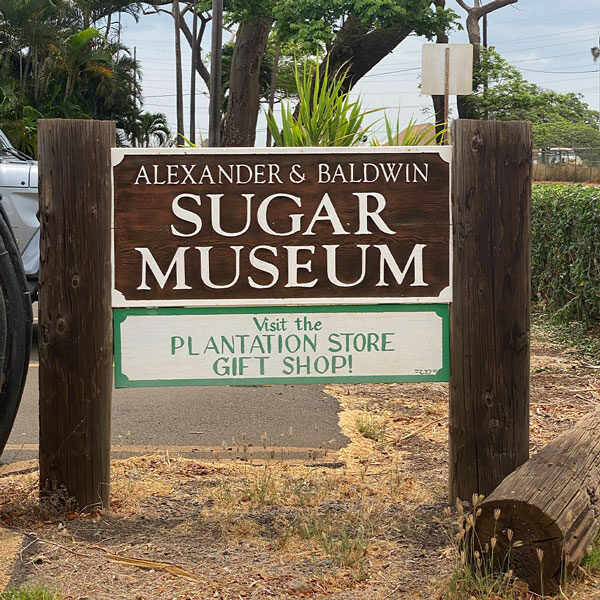 A wooden signboard reads, "Alexander & Baldwin Sugar Museum." Below, it says, "Visit the Plantation Store Gift Shop!" Surrounded by lush plants and trees, this scenic Maui spot invites you on a unique adventure steeped in history.