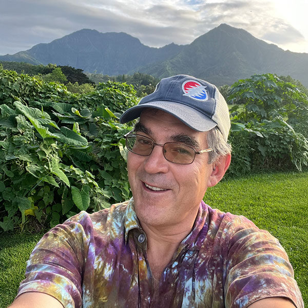 A person wearing a tie-dye shirt and a cap takes a selfie outdoors, capturing their joyful expression on an adventure in Maui. They're smiling in front of lush greenery and mountain peaks under a cloudy sky, perfectly embodying the spirit of exploration.